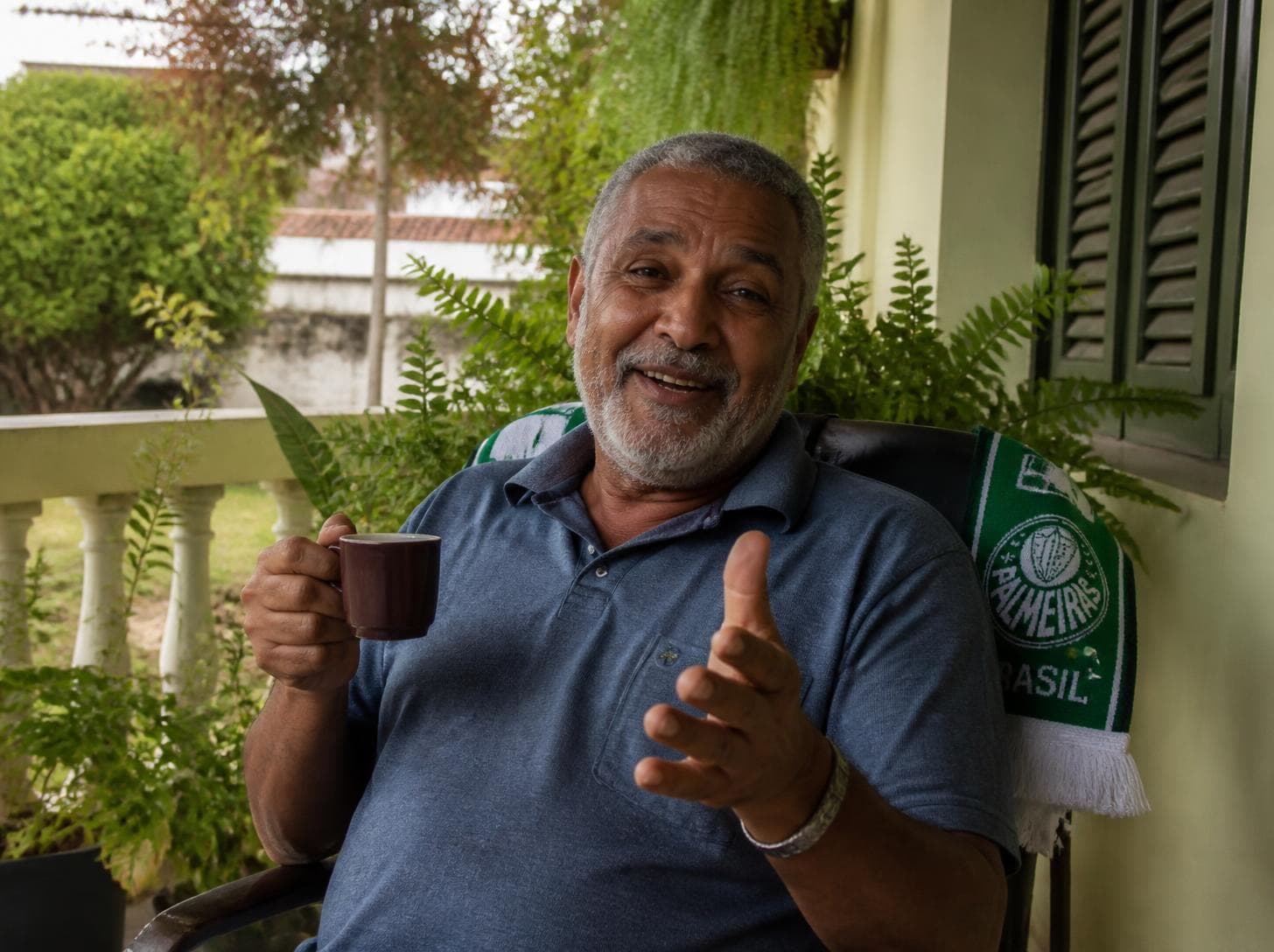Homem de barba grisalha contando uma história na varanda, com uma xícara de café na mão.